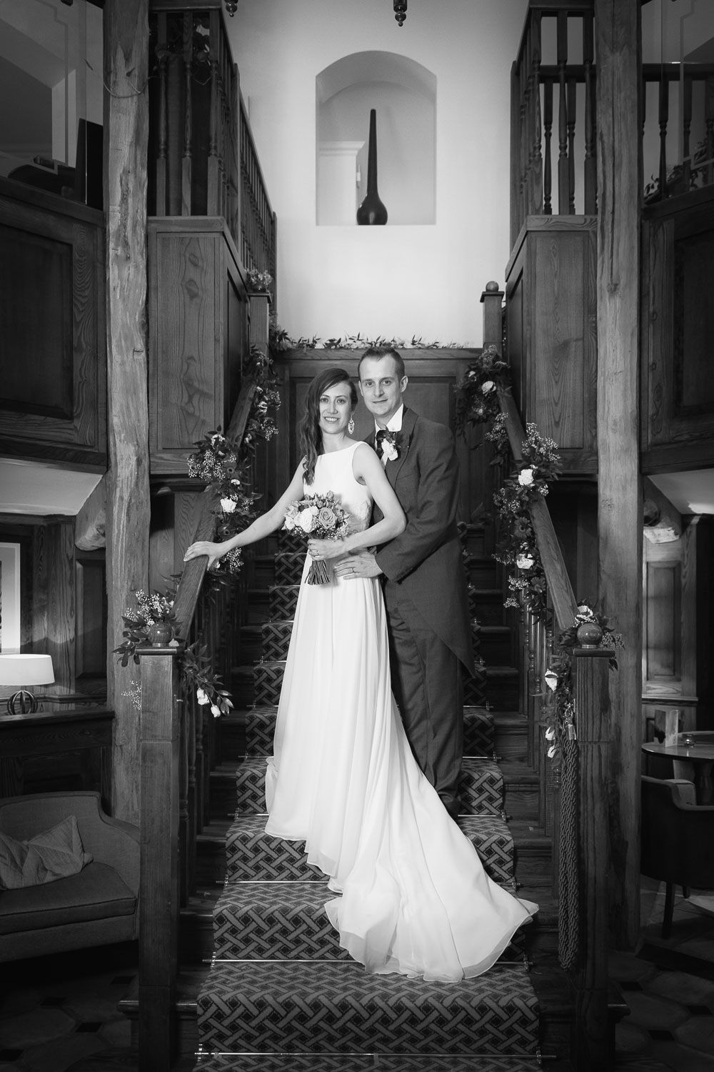 Black and white wedding photograph showing a formal couple posing on wooden church steps decorated with flowers.