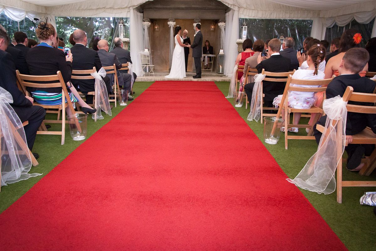Elegant outdoor wedding ceremony setup with red carpet aisle runner and wooden chairs on grass lawn.