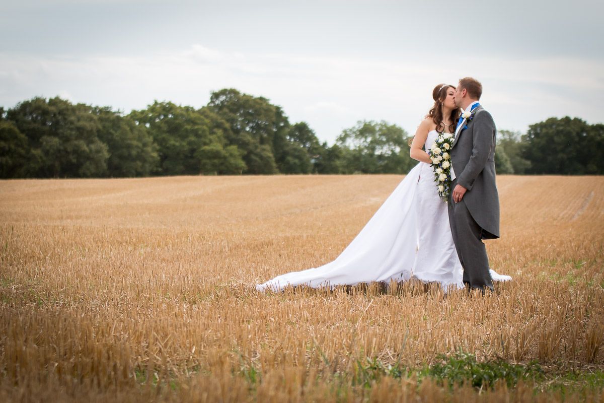 A bride and groom embrace in a serene wheat field on their wedding day against a cloudy sky backdrop.