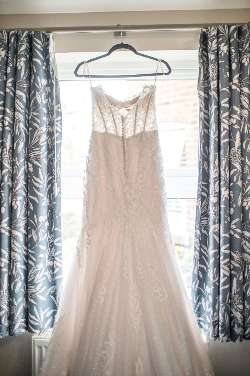 Multiple elegant white wedding dresses hanging on hangers in front of a window with blue floral curtains.