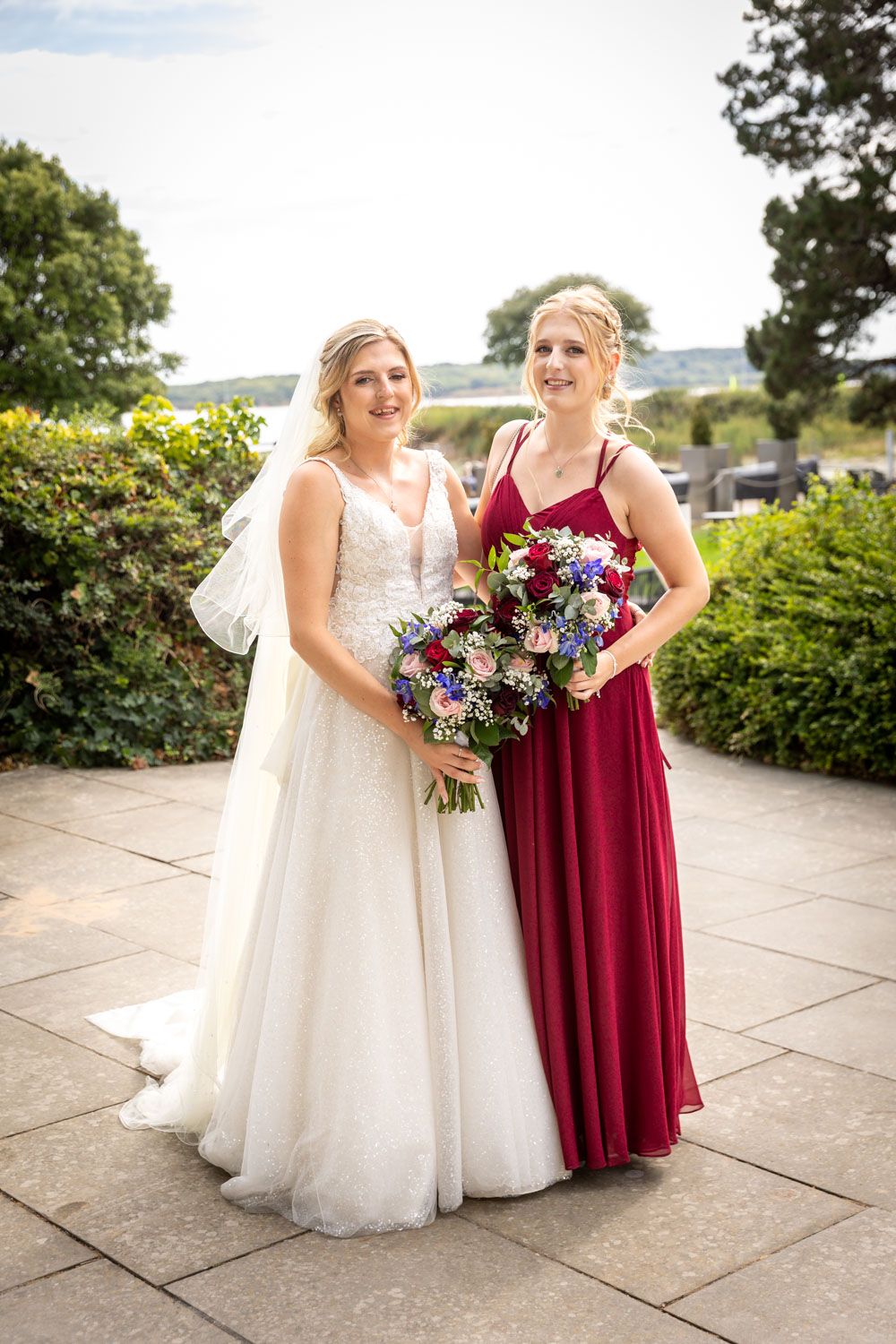 Bride in white lace gown and bridesmaid in red dress pose together holding colorful bouquets outdoors.