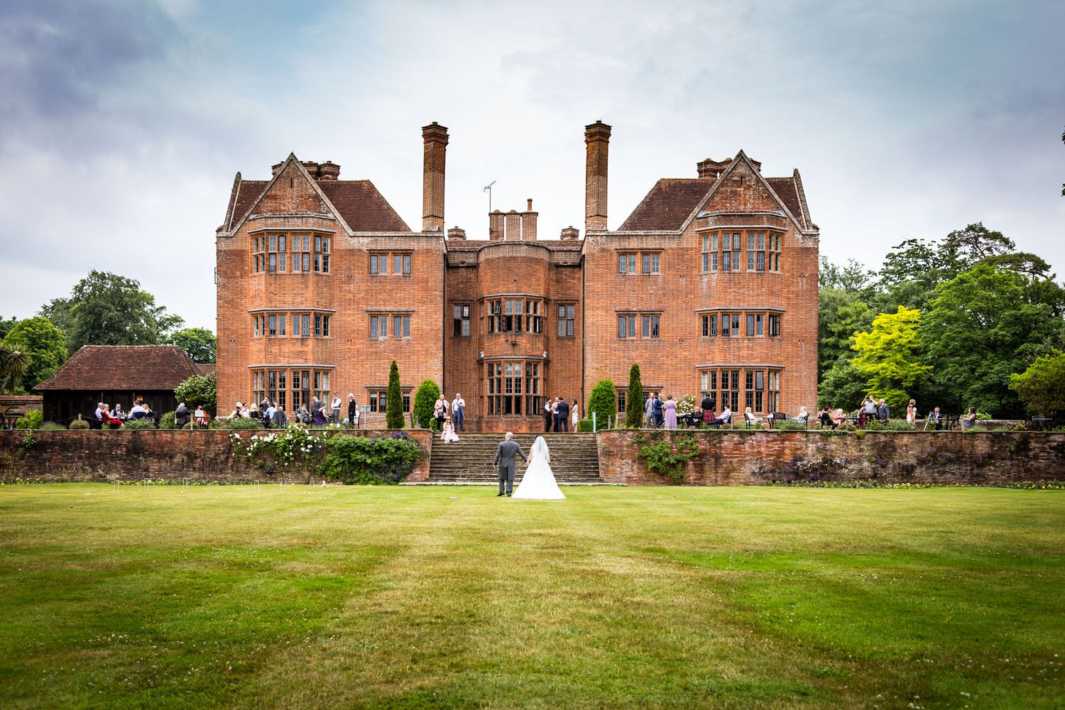 Wedding couple stands on vast lawn before elegant red-brick Tudor manor.