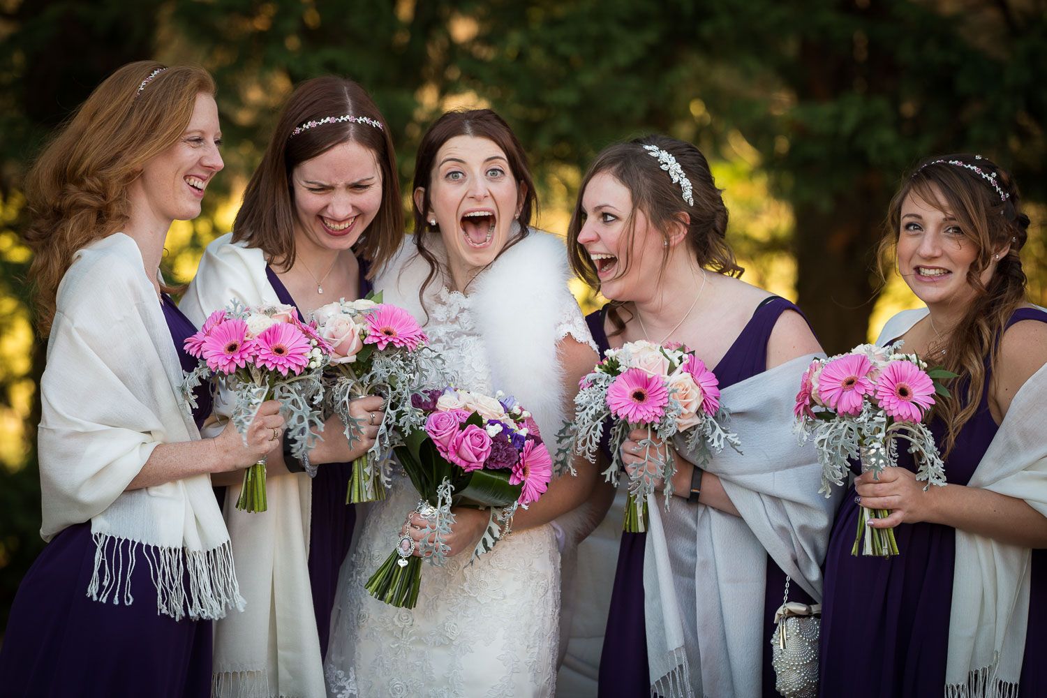A bridal party holding pink flower bouquets shares a joyful candid moment together outdoors.