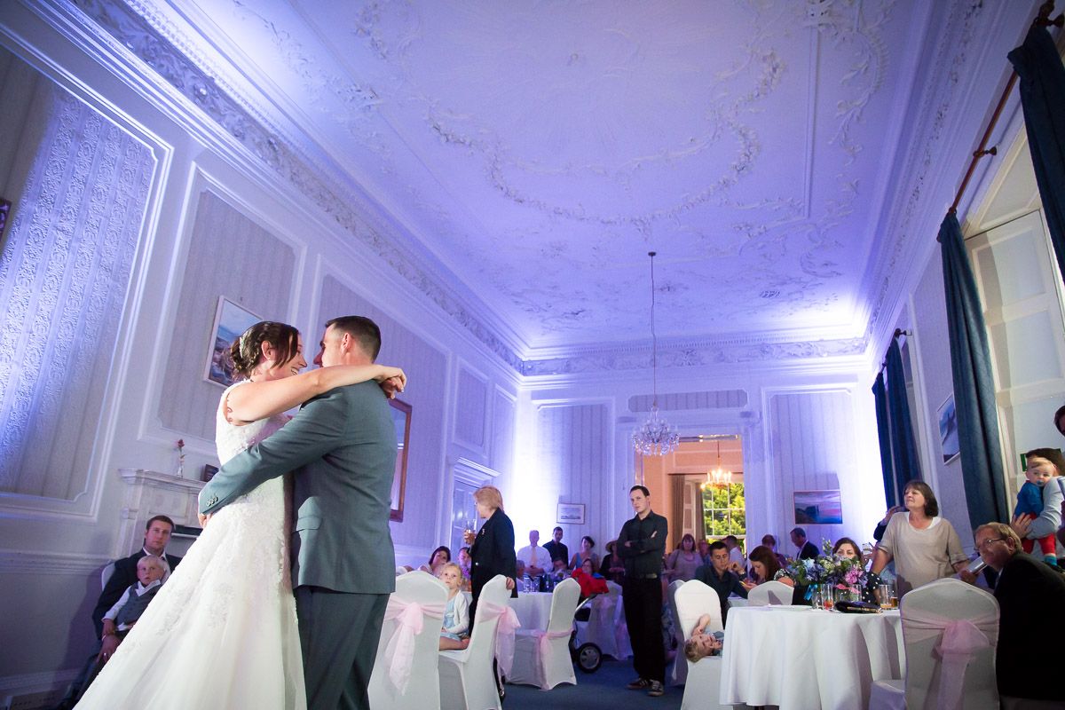 A couple shares their first dance in an elegant blue-lit ballroom surrounded by seated wedding guests.