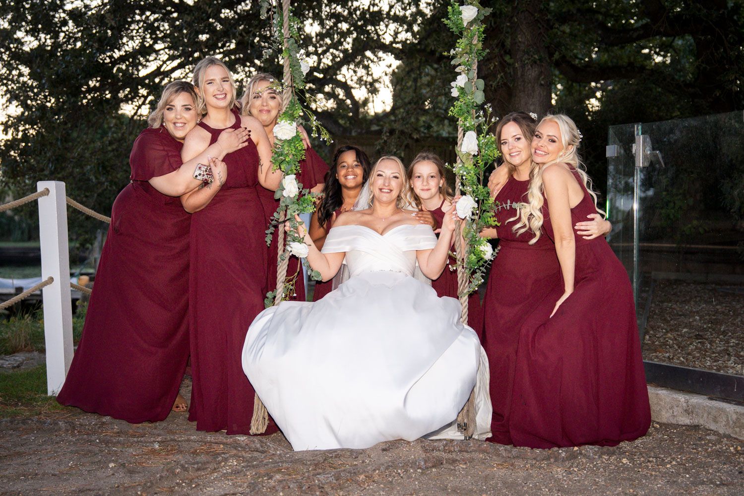 Wedding party in burgundy dresses poses together for a series of group photos on the venue grounds.