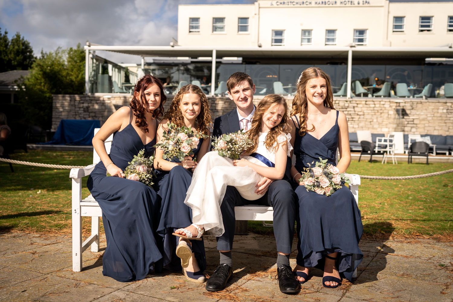 Wedding party members in navy blue dresses pose together on outdoor bench during sunny celebration.