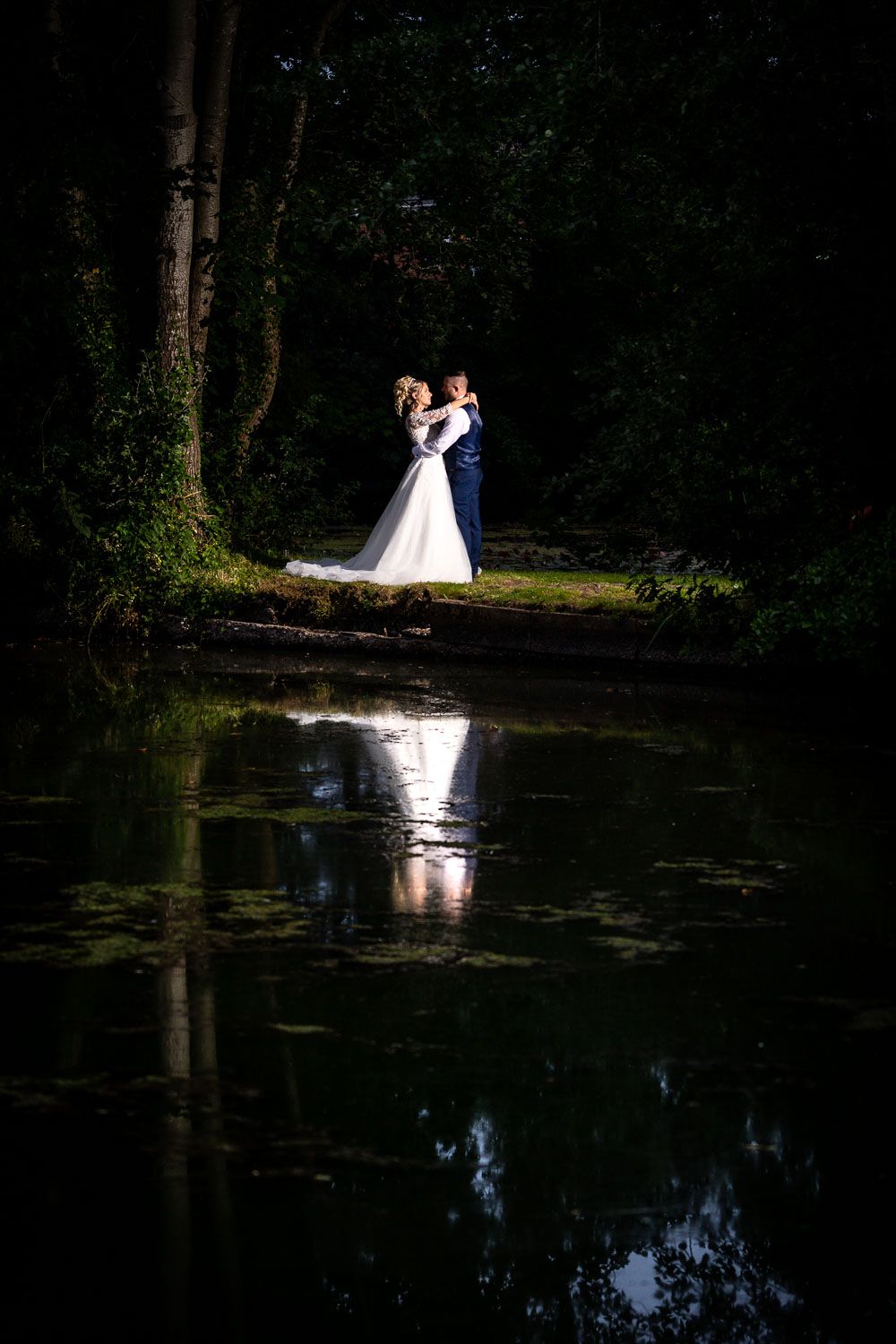 A couple shares a romantic woodland moment beside a tranquil pond that perfectly mirrors their embrace in its dark waters.