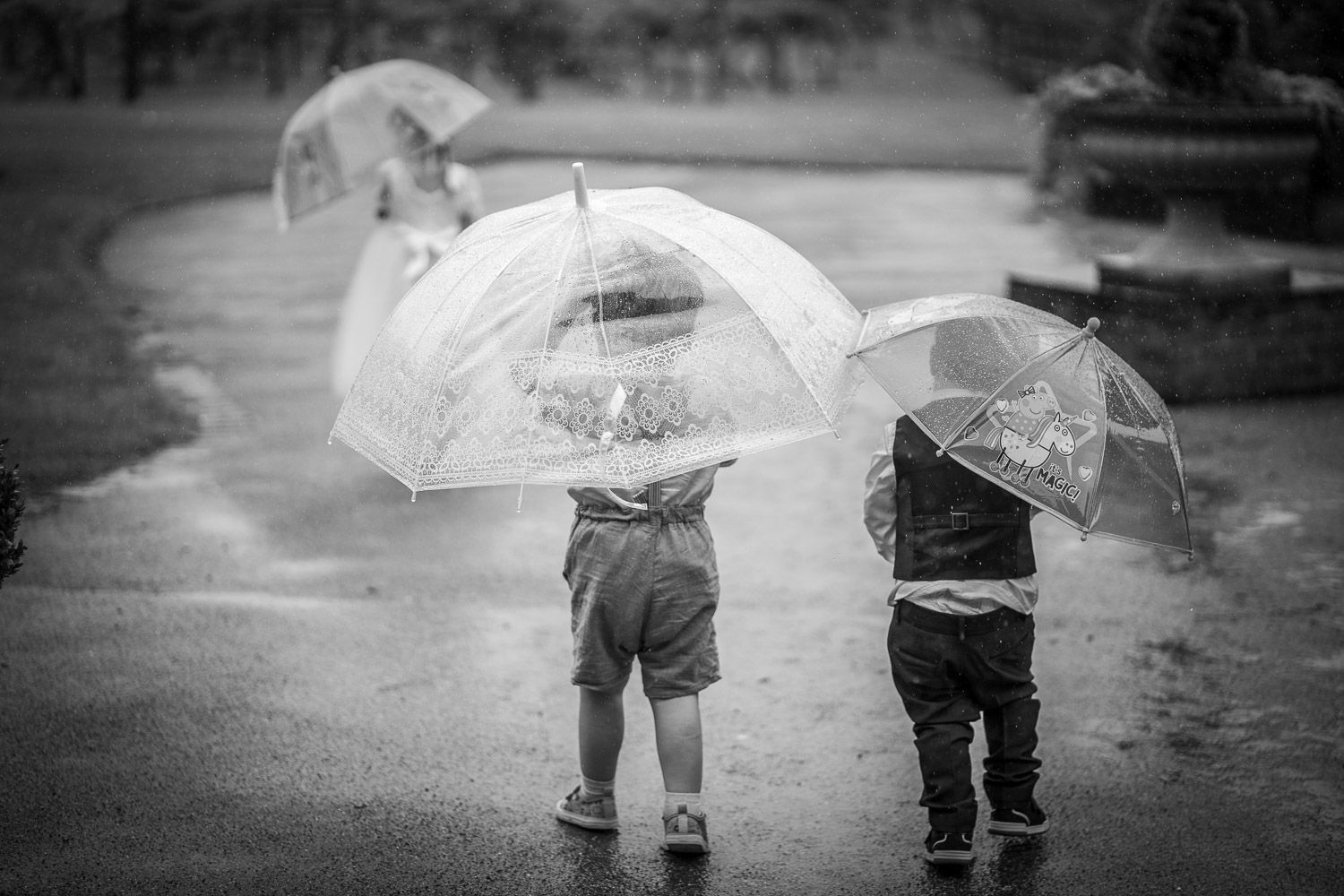Two small children hold transparent umbrellas while standing on a wet sidewalk in black and white photography.