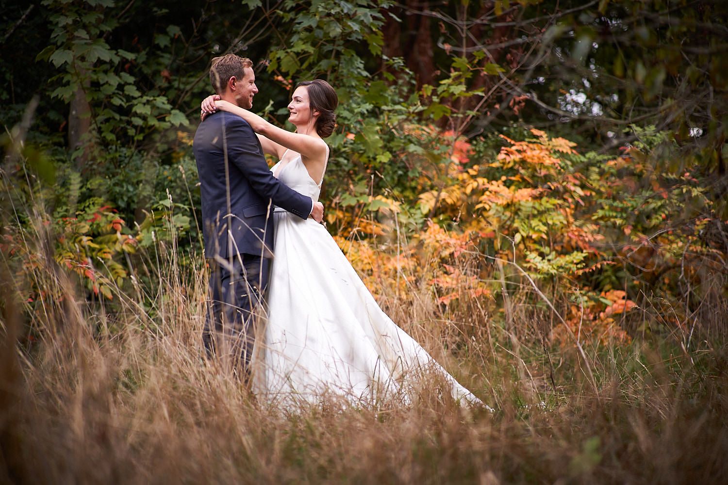 A couple in wedding attire dance together in a fall meadow with colorful autumn foliage in the background.