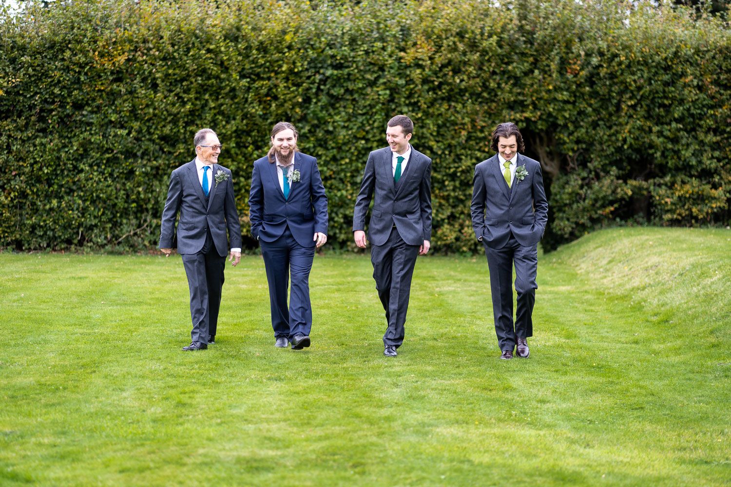 Four groomsmen in navy blue suits walk together across a lush green lawn with a tall hedge backdrop.
