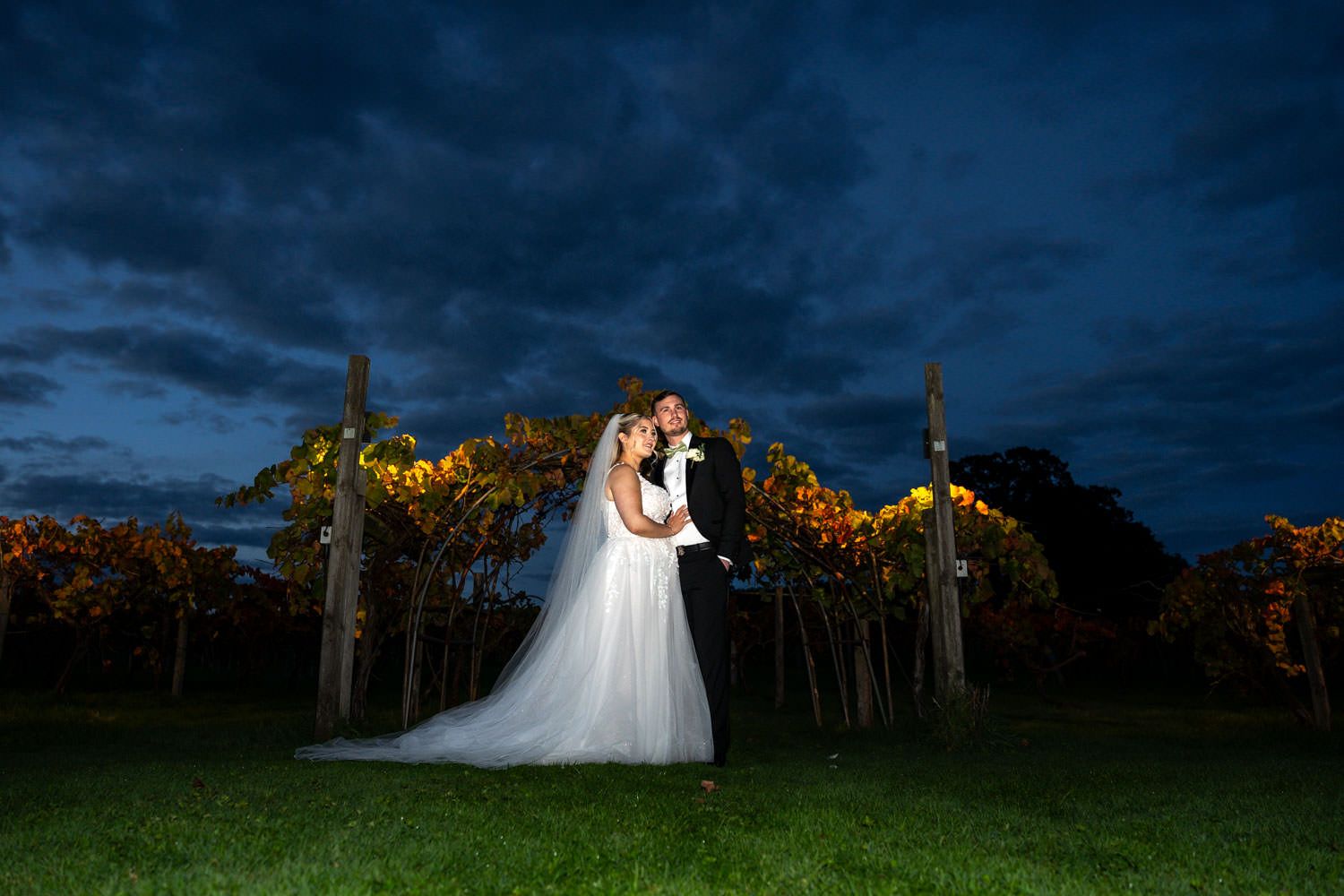 Bride and groom share a romantic embrace in an autumn vineyard under a dramatic twilight sky.