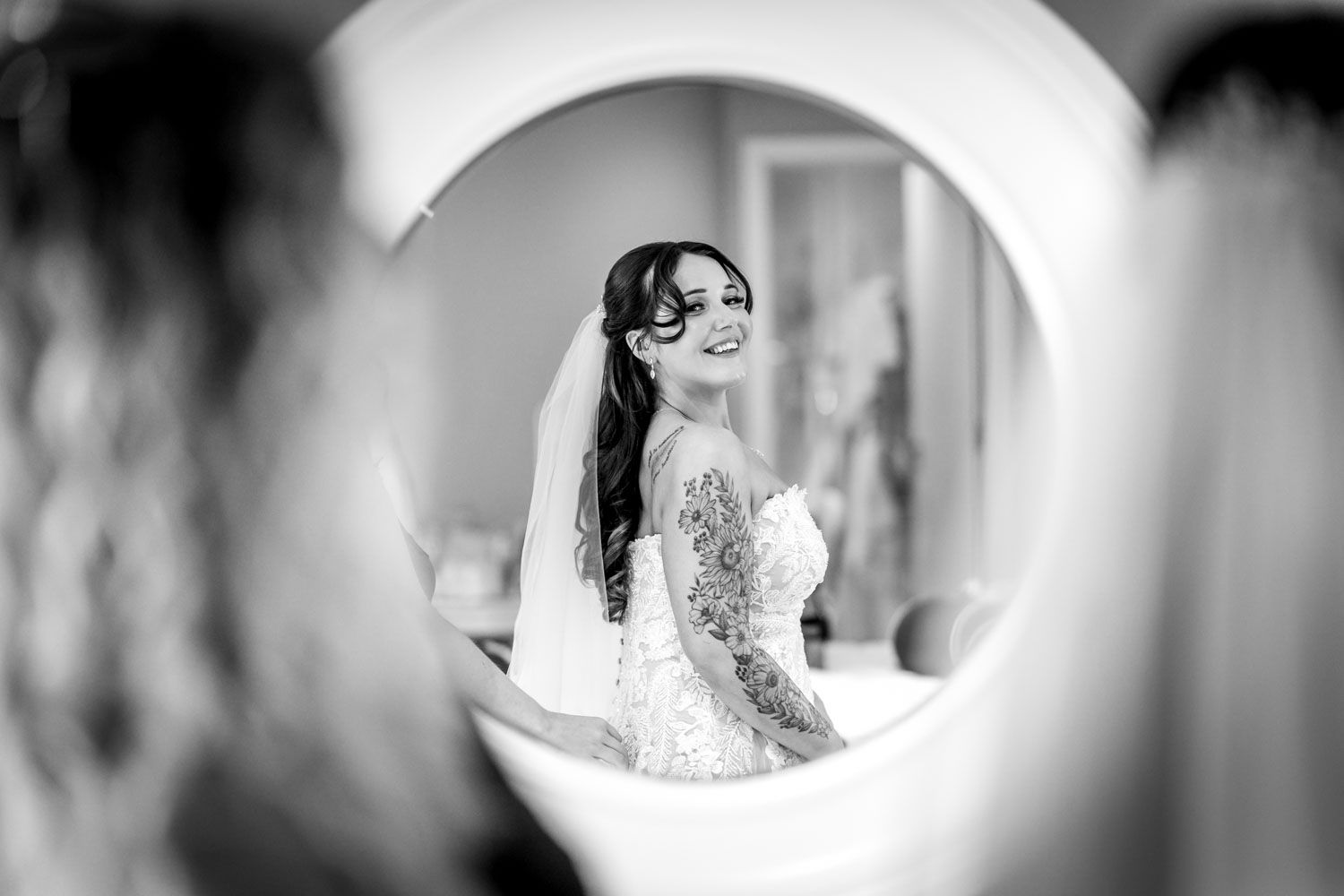 Black and white mirror reflection of smiling bride in lace wedding dress during preparations.