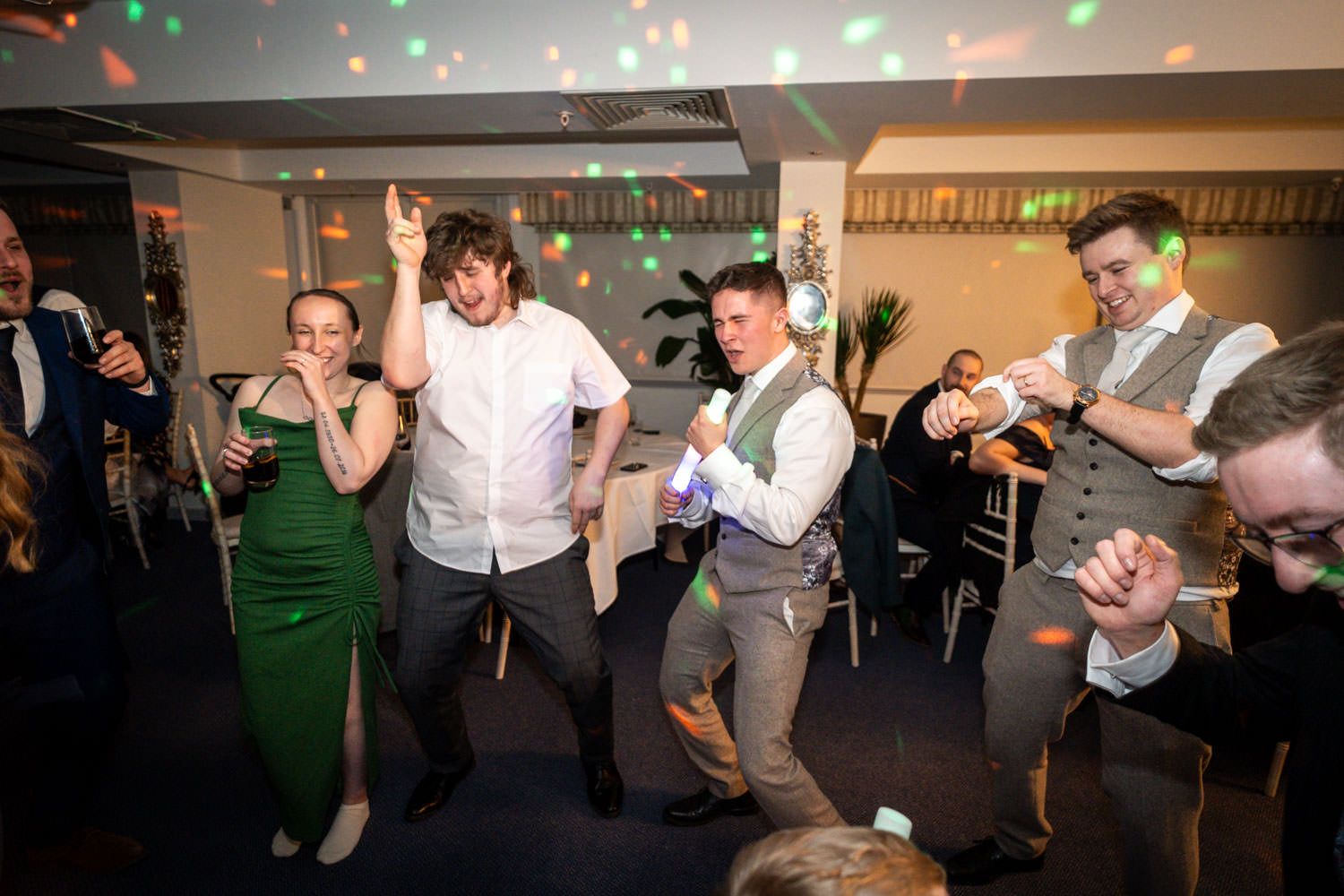 Party guests dance and celebrate on the dance floor at an indoor event with festive lights overhead.