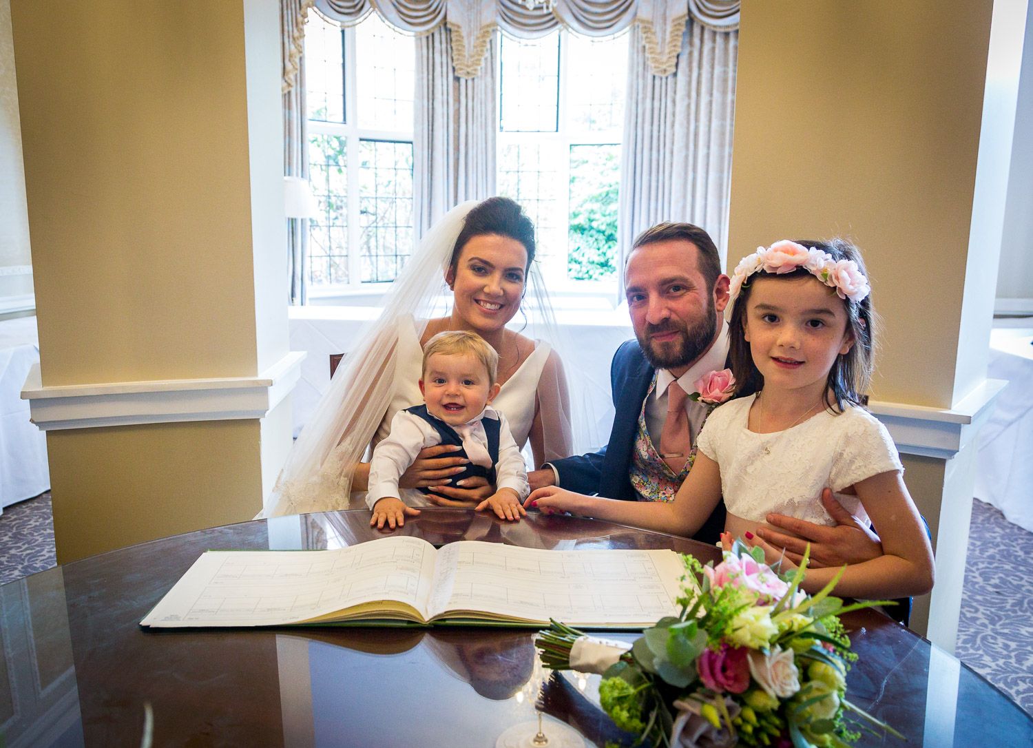 Family gathers around a book at a table with floral decorations near elegant draped windows.