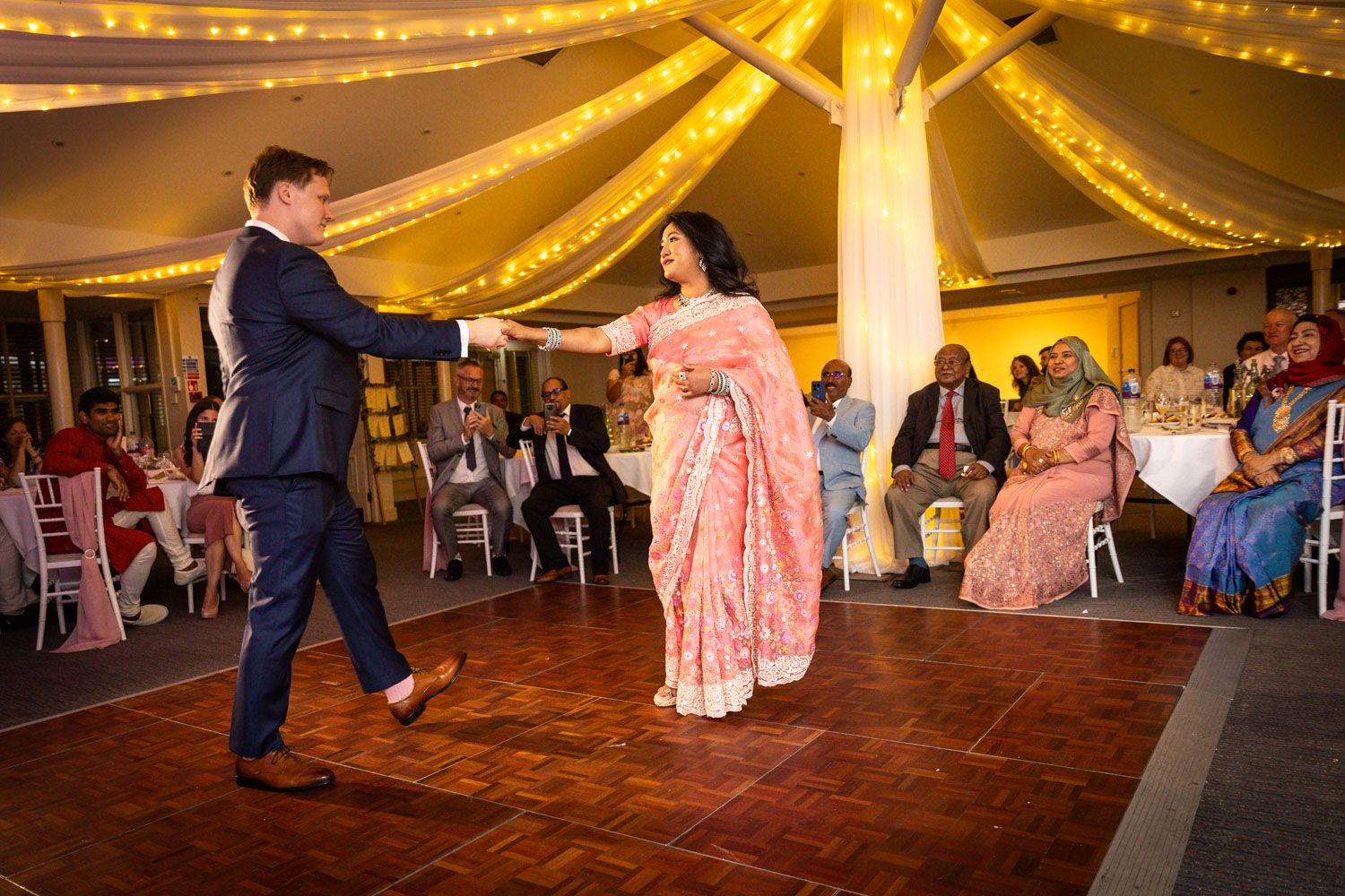 A couple dances on a wooden floor under a tent decorated with warm glowing string lights at an elegant reception.
