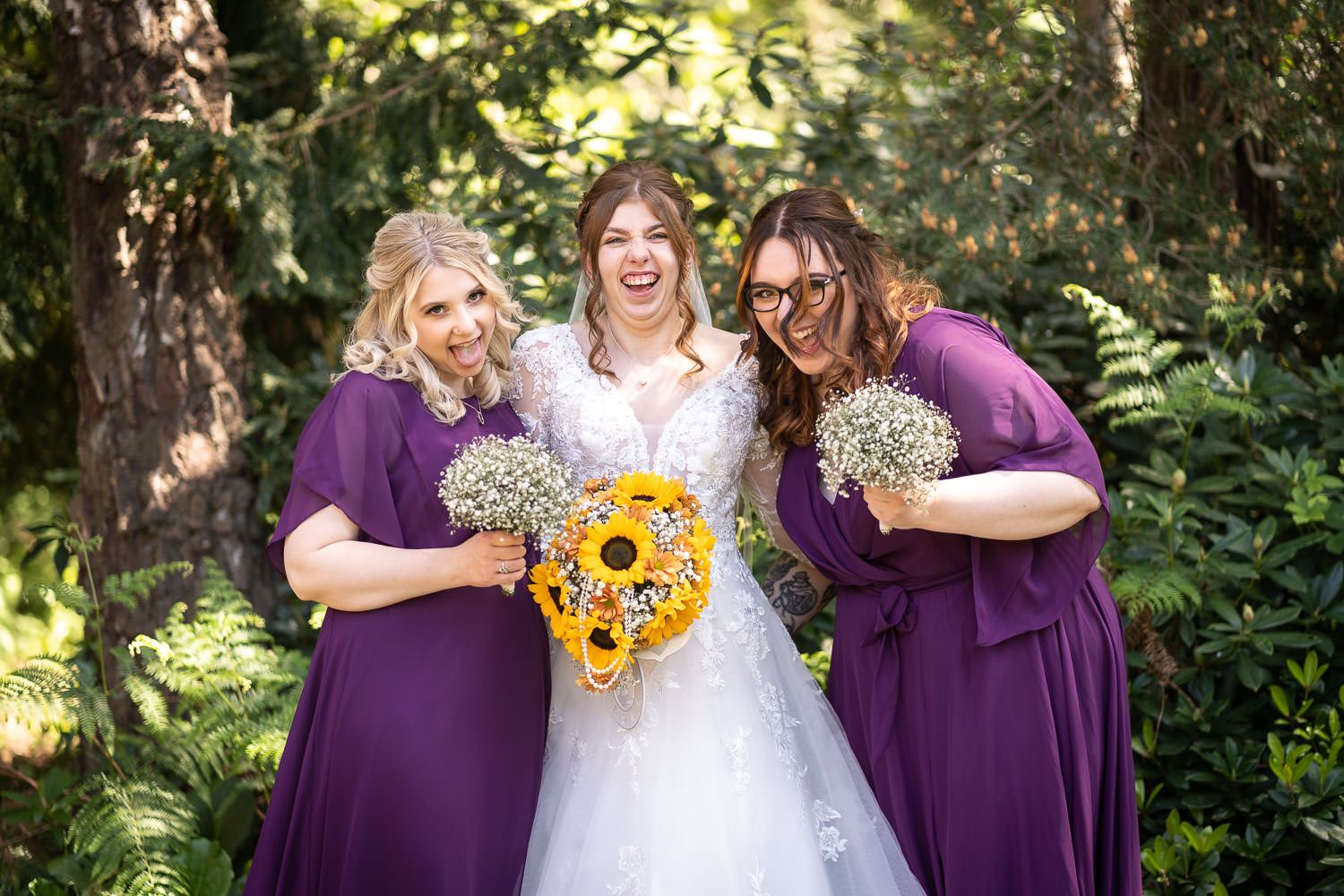 Three women in white and purple dresses share a joyful moment while holding bouquets of sunflowers and wildflowers outdoors.