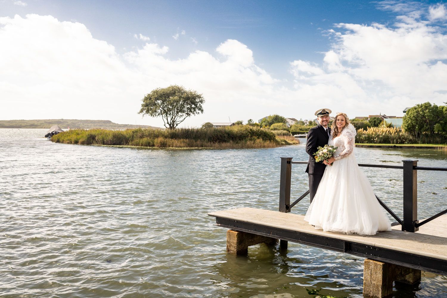 A couple stands on a wooden dock overlooking the water during their wedding ceremony on a sunny day with blue skies.