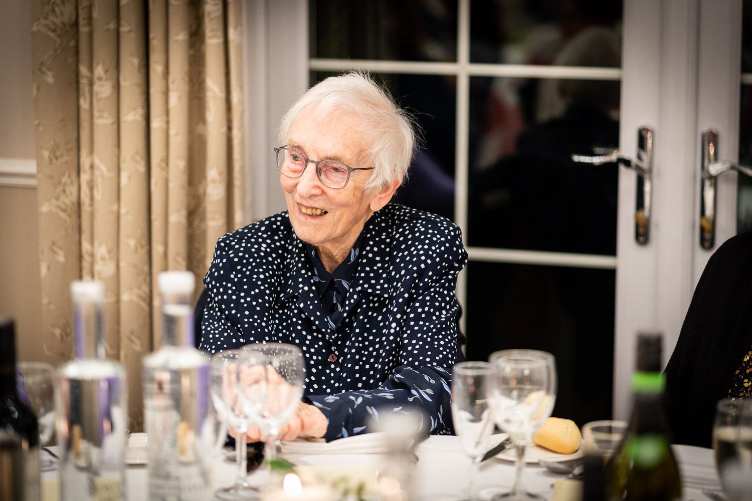 An elderly person in a polka dot shirt smiles warmly during a meal at a dining table with glassware.