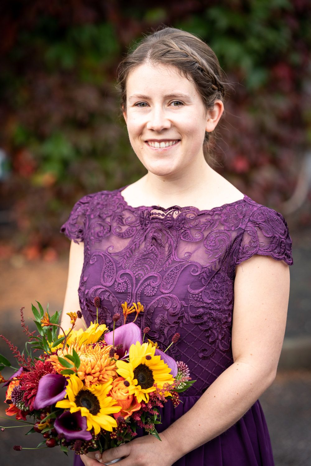 Someone wearing a purple lace dress holds a vibrant fall bouquet with sunflowers and purple calla lilies.