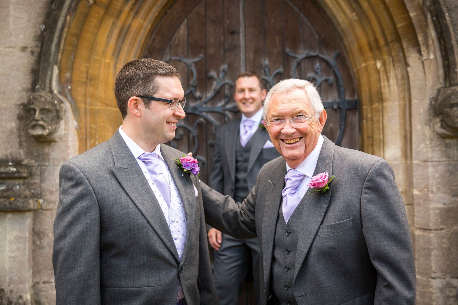Three people in formal wedding attire share a candid moment in front of a stone archway.
