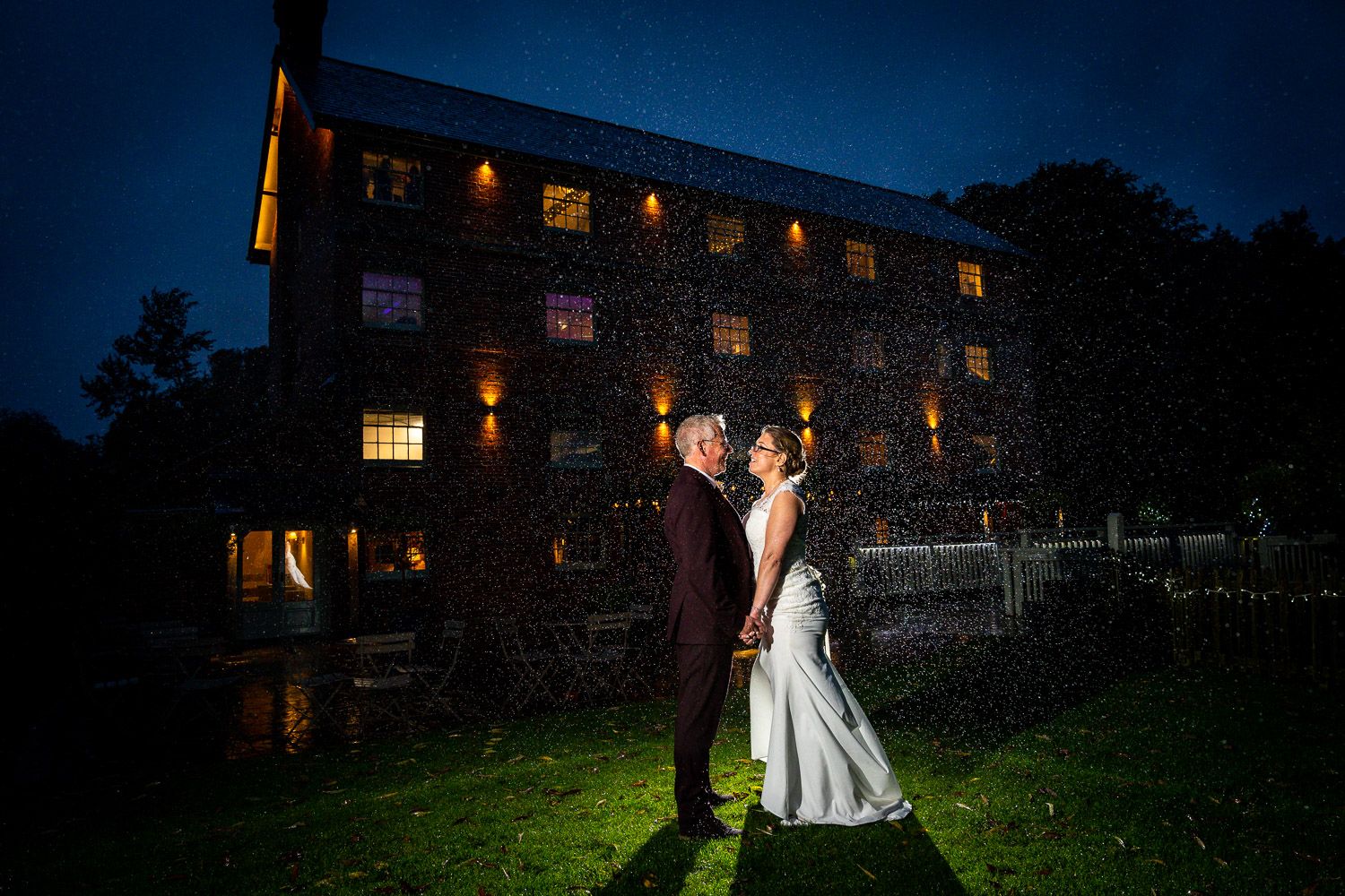 A romantic nighttime wedding portrait in front of an illuminated stone mill with glowing windows.