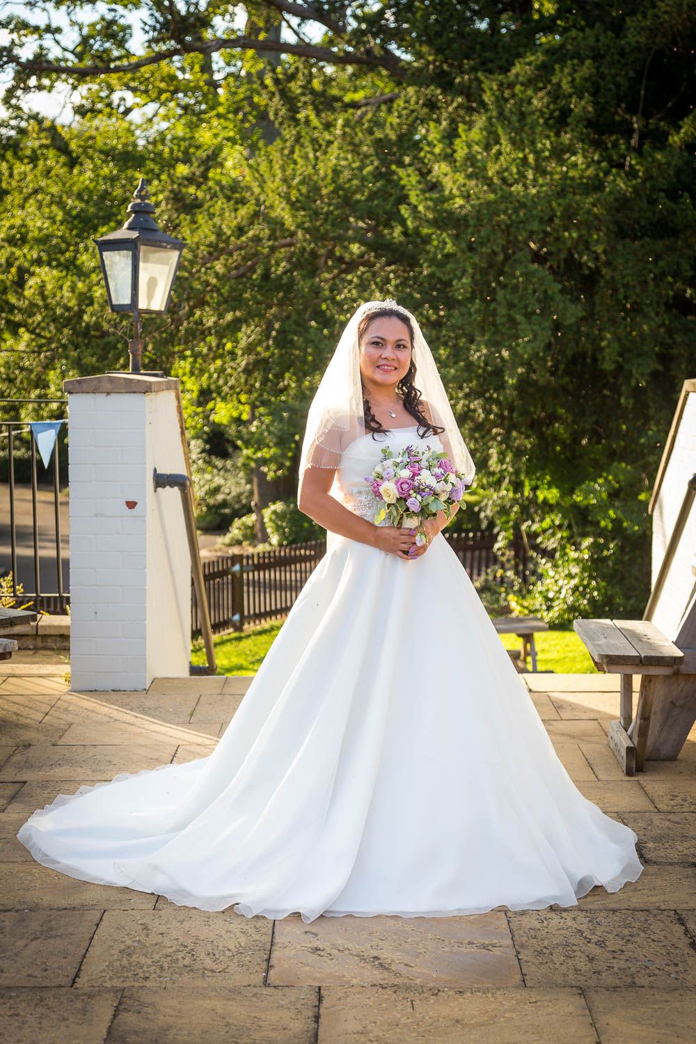 A bride in a white ballgown wedding dress holds a pink and purple bouquet on a stone patio with decorative lamp post.