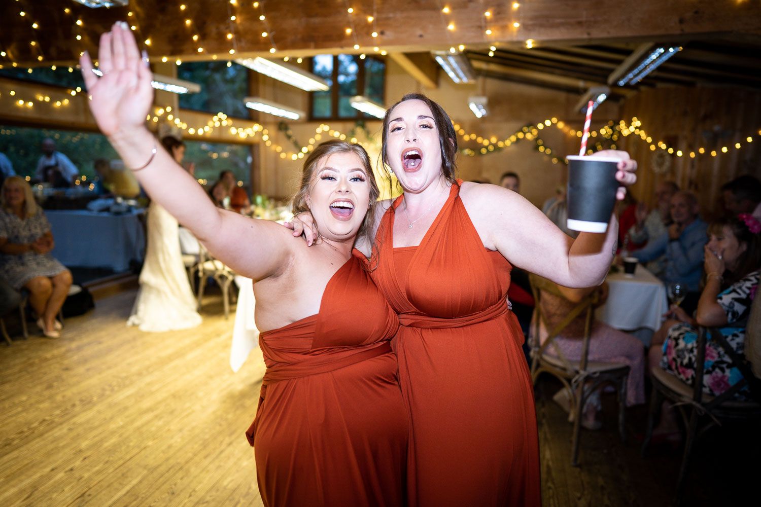Two people in matching rust-colored dresses dancing together at a reception venue decorated with string lights.