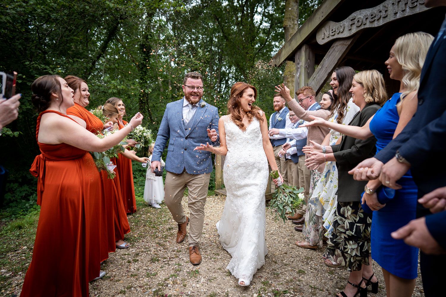 Joyful wedding couple walks through path lined with guests throwing flower petals at outdoor woodland ceremony.