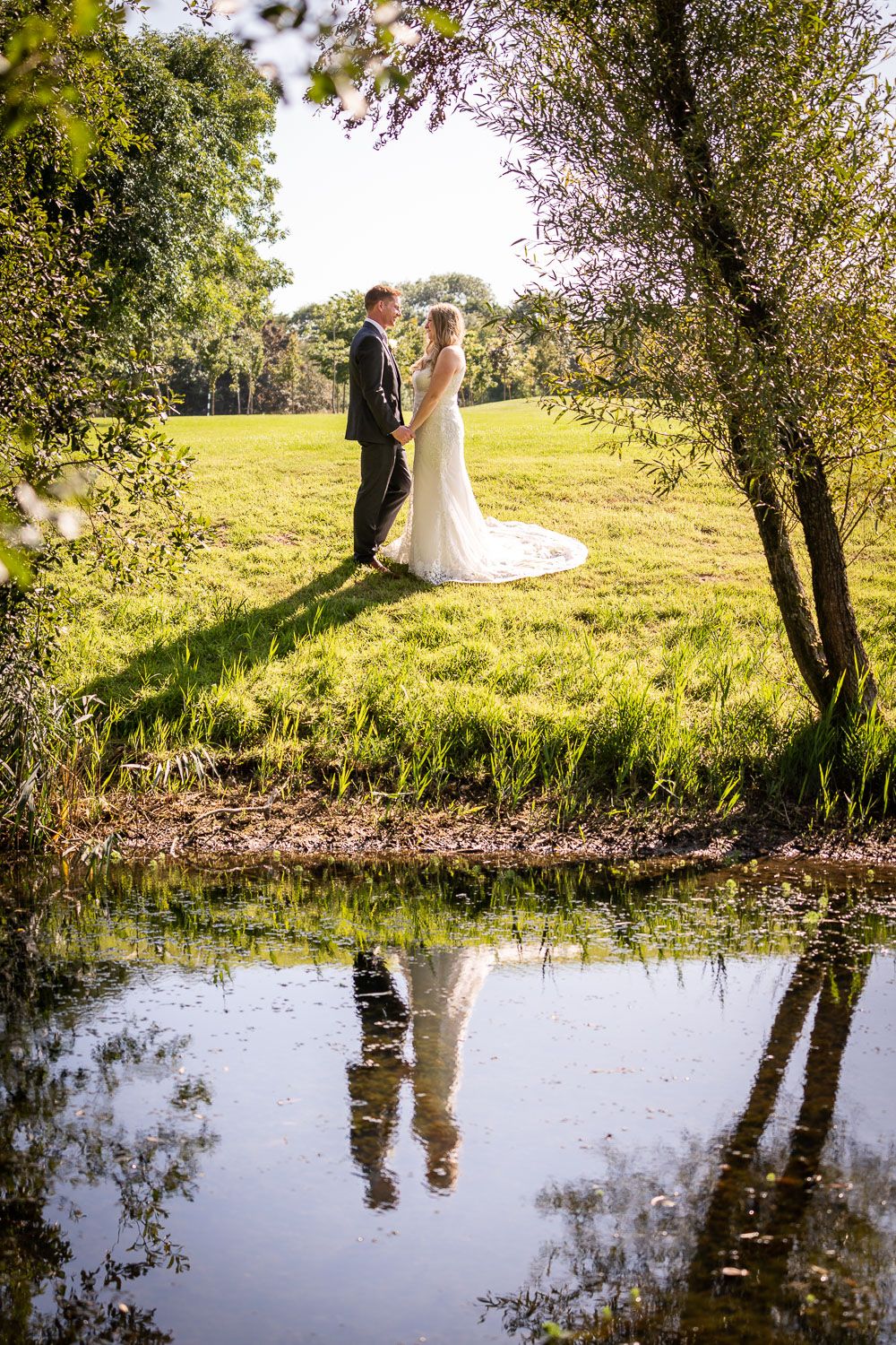 A wedding couple stands near a pond on a sunny day, their reflection mirrored in the still water beneath blossoming trees.