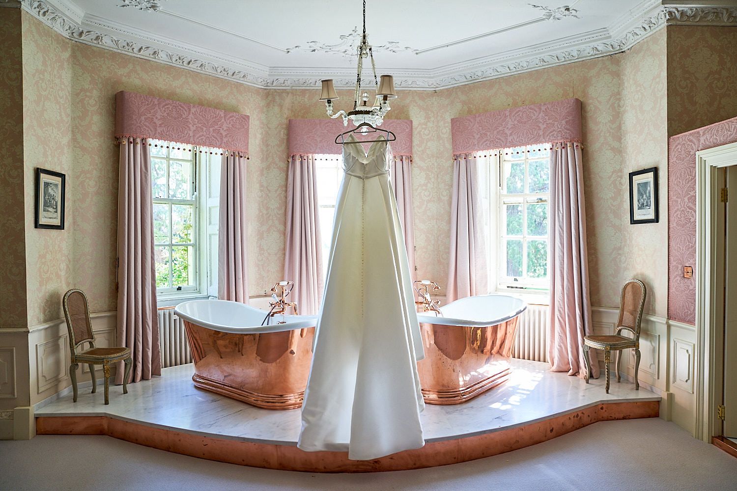 Elegant bridal gown hangs in luxurious bathroom with copper bathtubs and pink walls decorated with ornate crown molding.