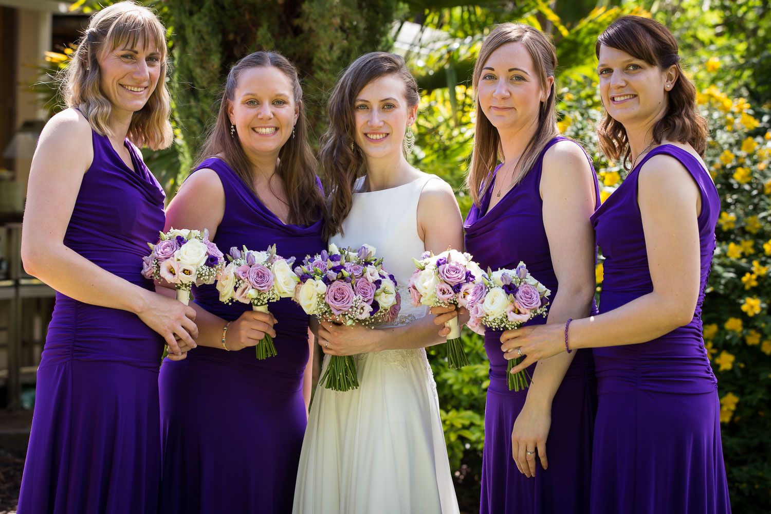 Wedding party poses outdoors in coordinated purple bridesmaid dresses holding pink and purple floral bouquets.