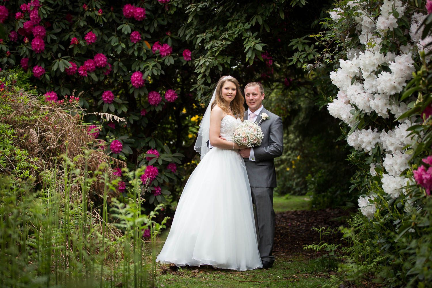 A wedding couple stands in a garden path surrounded by blooming pink and white flowers in full summer bloom.