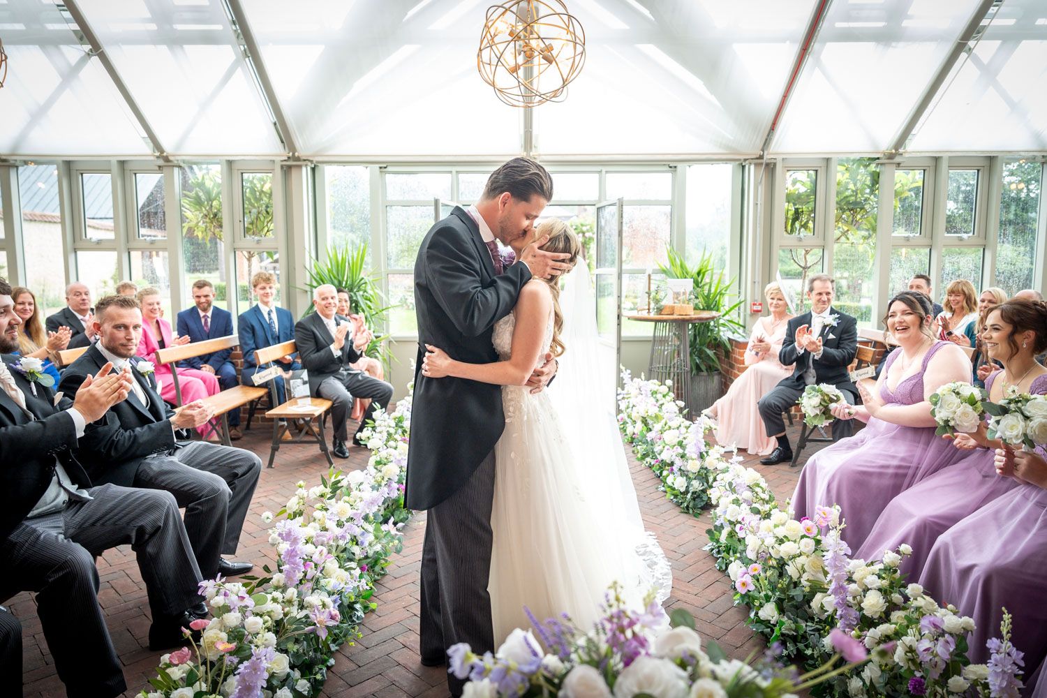 Wedding ceremony in a bright conservatory filled with floral arrangements and seated guests.