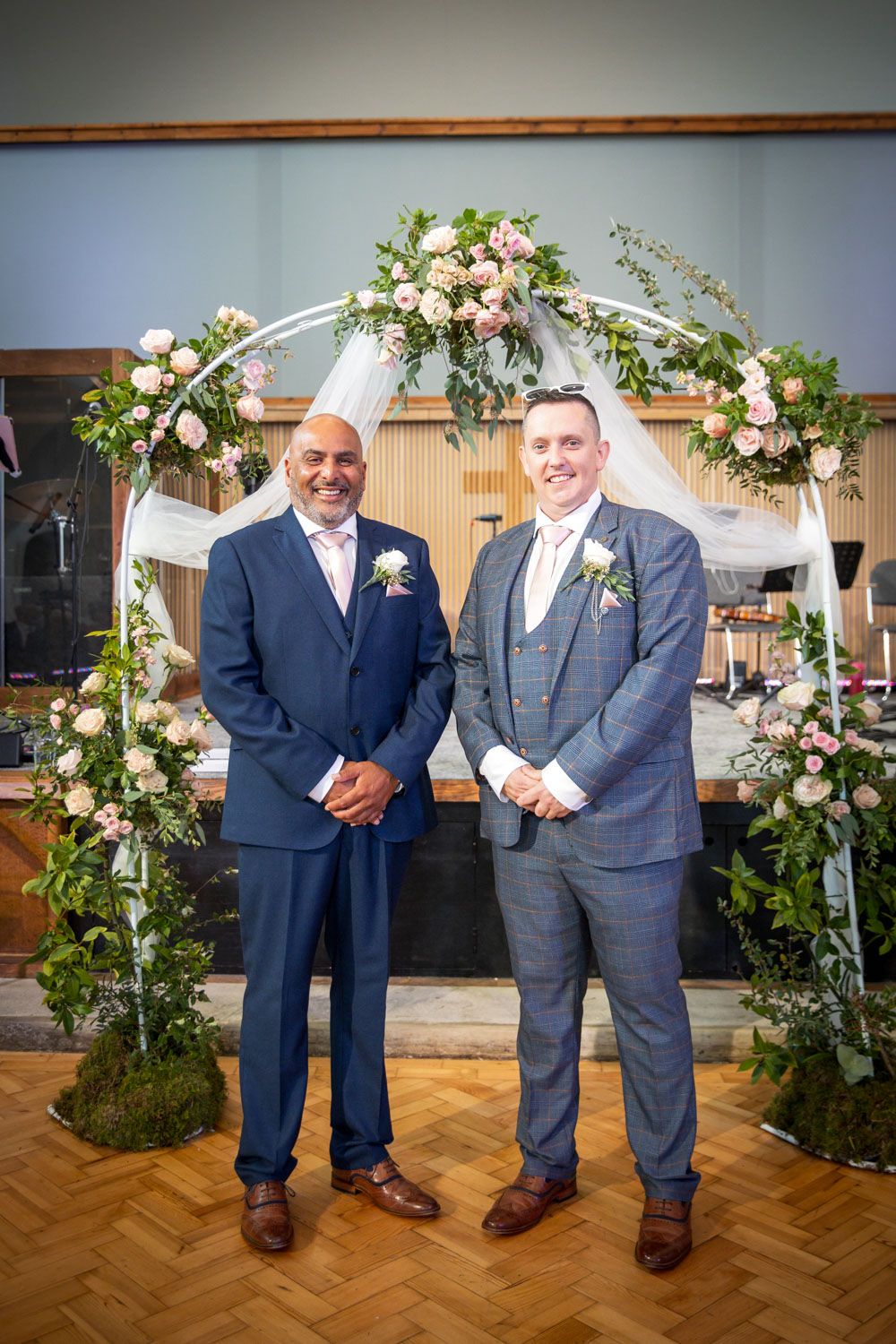 Two people stand together in formal navy suits near a decorative floral arch with roses at an indoor wedding venue.
