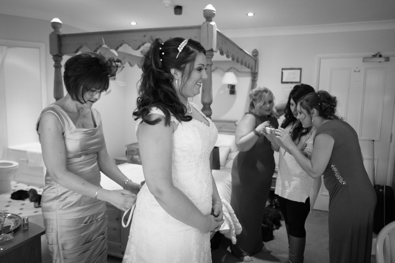 A candid black and white photo showing wedding day preparations with people getting ready in a bedroom setting.