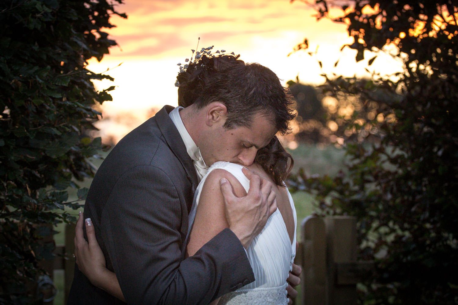 Bride and groom share an intimate embrace at sunset framed by green hedges at their wedding.