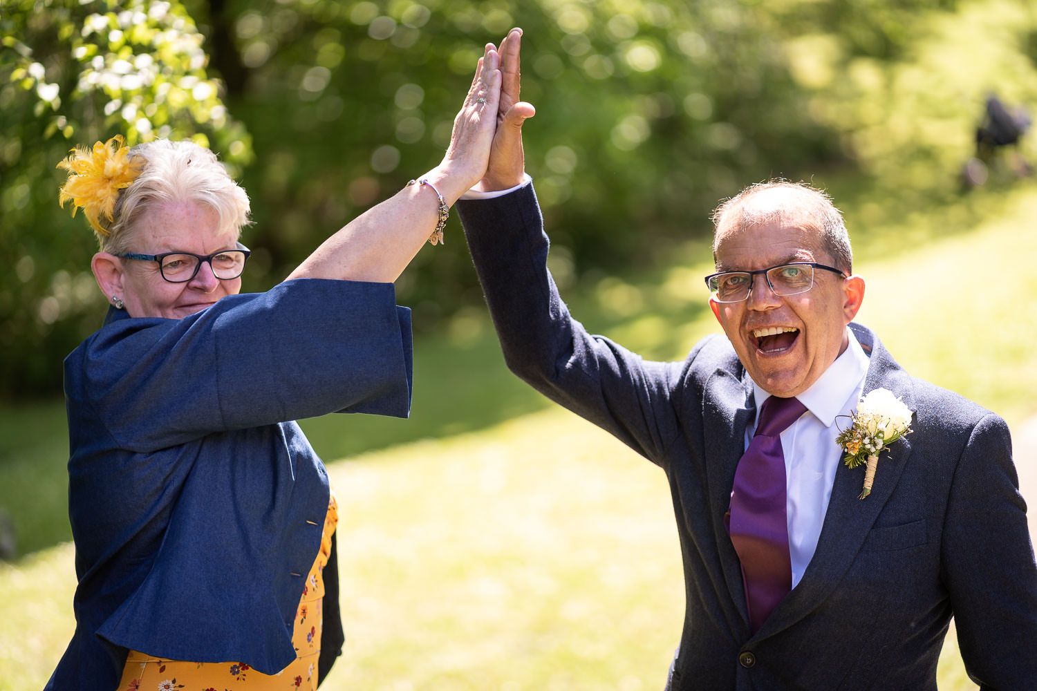 Two joyful seniors in formal attire give each other an energetic high five outdoors on a sunny day.