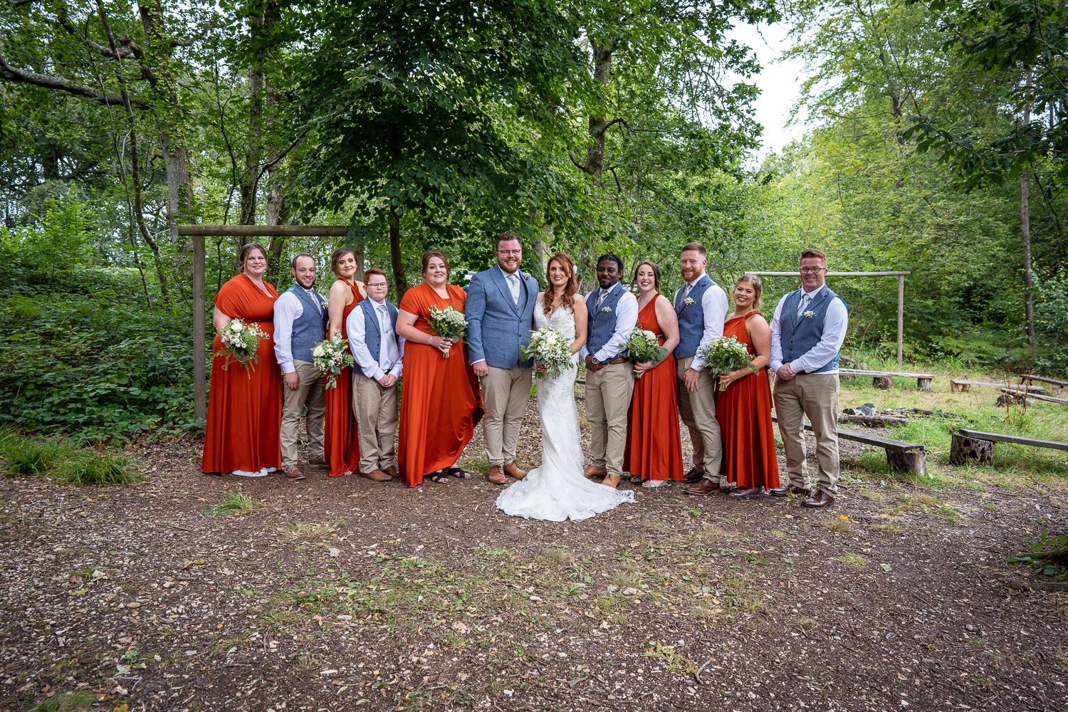 Wedding party in rustic outdoor setting wearing coordinating red dresses and gray suits pose together in woodland area.