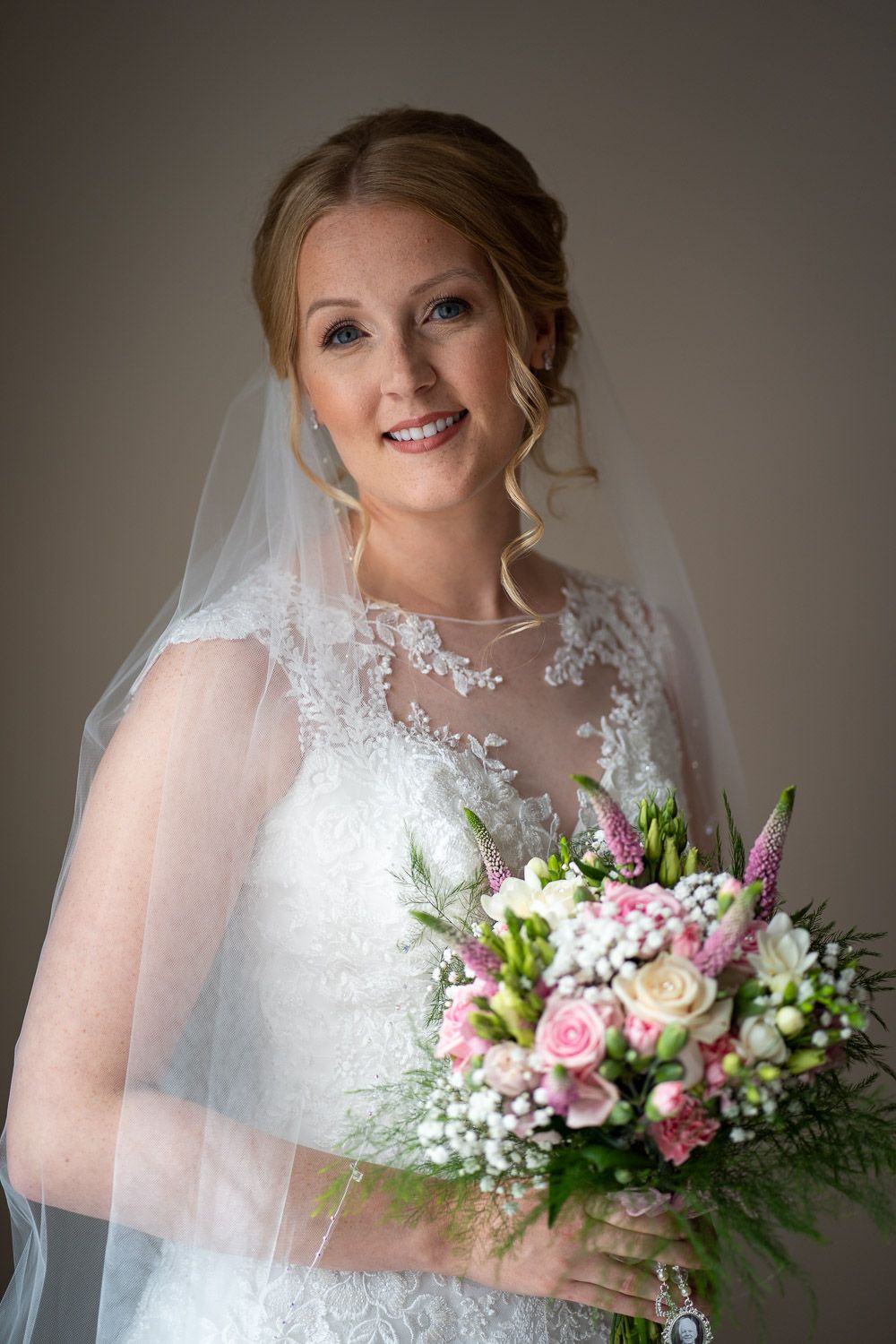 A bride in a delicate lace dress with sheer overlay holds a beautiful bouquet of pink roses and white flowers.