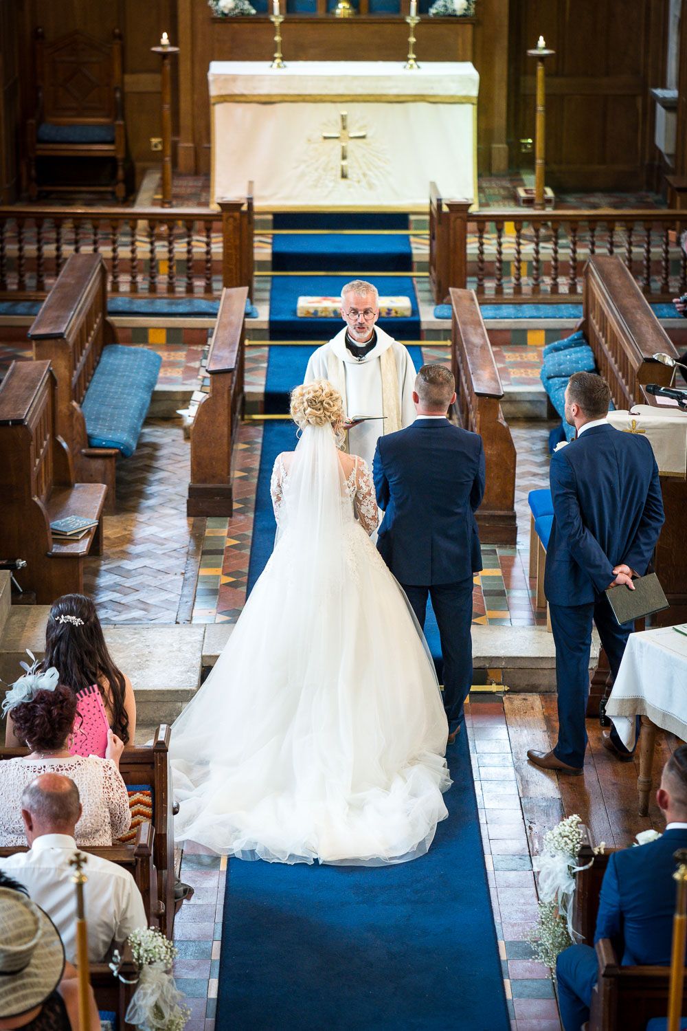 Traditional church wedding ceremony with couple standing at altar on blue carpet aisle runner surrounded by wooden pews.