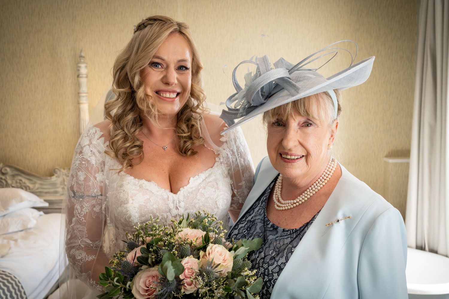 A bride in a white lace wedding dress poses with an older woman wearing a light blue outfit and fancy hat.