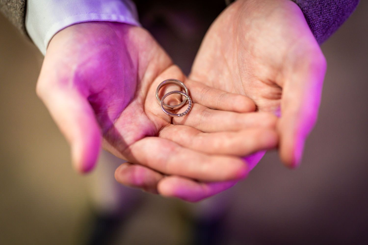 Two hands holding shiny wedding rings in cupped palms illuminated with purple lighting.