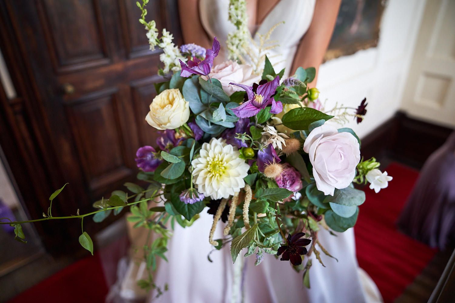 A beautiful wedding bouquet featuring purple, white, and blush flowers with green eucalyptus leaves held against a white dress.