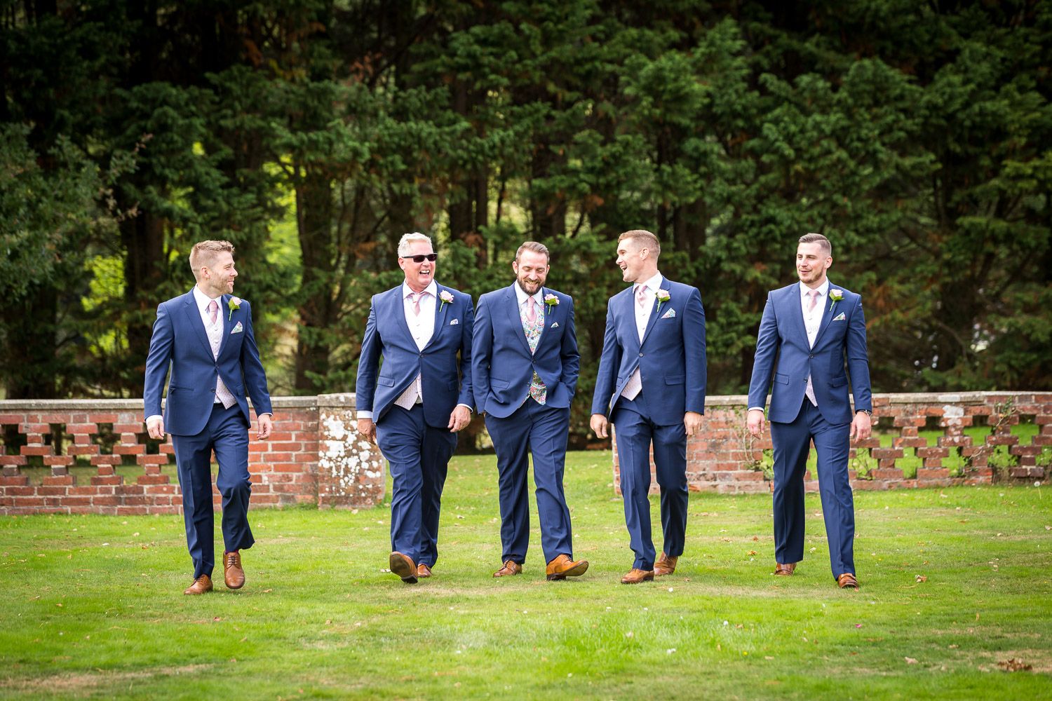 A group of five men wearing matching navy blue suits walking together on a green lawn near a brick wall.
