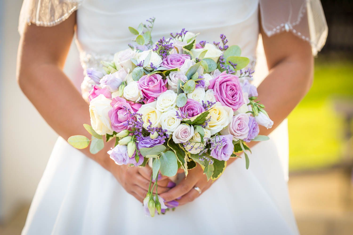 Beautiful bridal bouquet featuring soft pink roses, white flowers, and purple blooms held against a white dress.