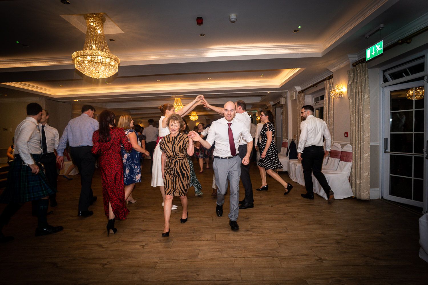 Guests dancing together at an elegant wedding reception with a crystal chandelier overhead on hardwood floors.