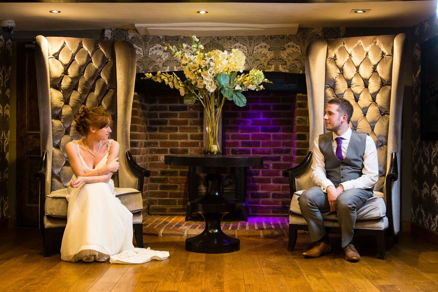 Two people seated in elegant padded alcoves with brick wall and floral arrangement between them in an atmospheric venue.