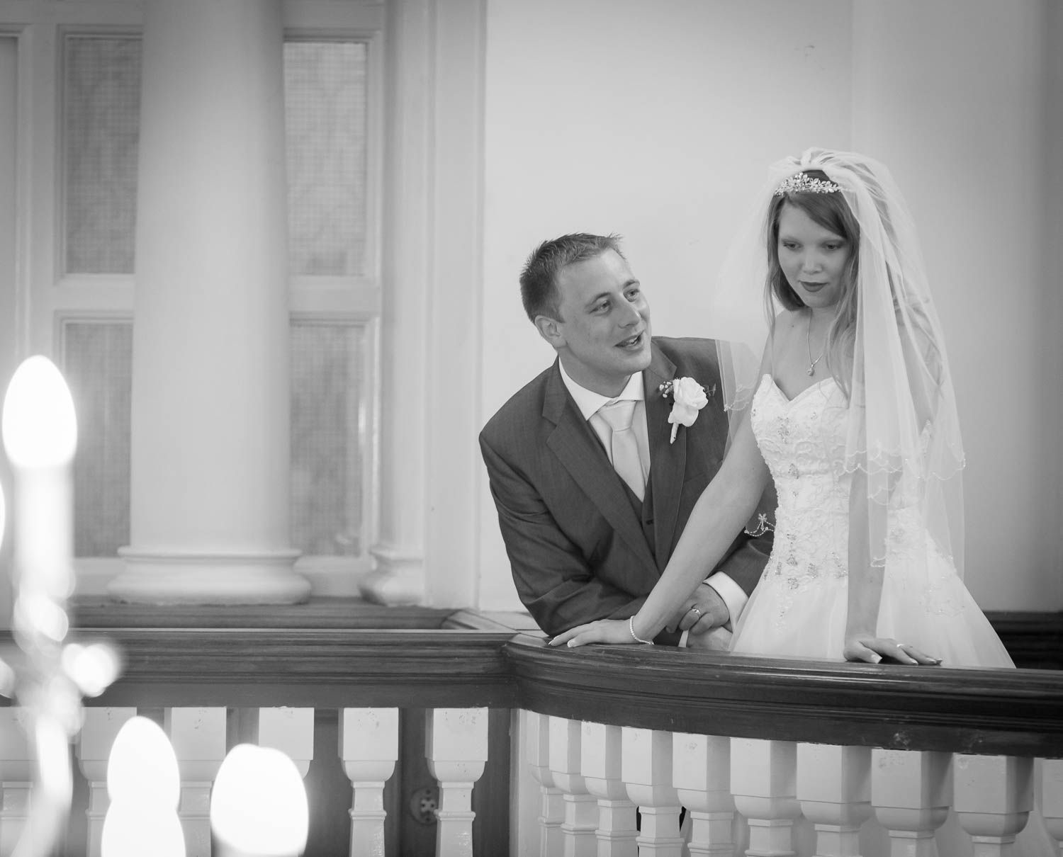 Black and white wedding photograph showing a couple on a balcony with architectural details in classic formal attire.