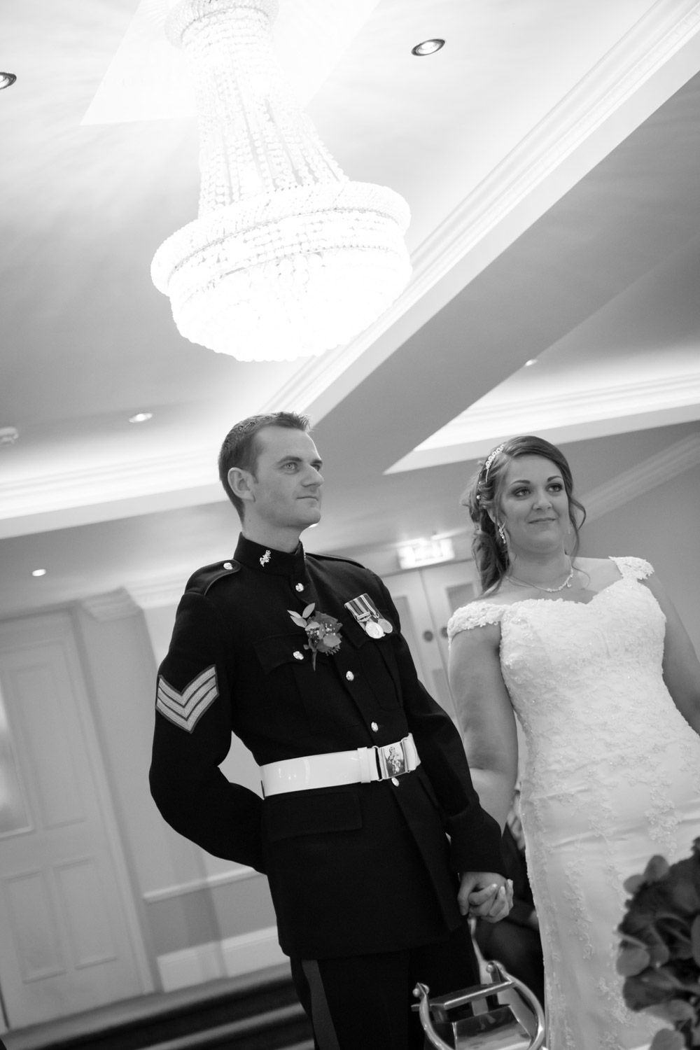 Black and white photo of a military wedding ceremony showing a uniformed serviceman and bride under an elegant chandelier.