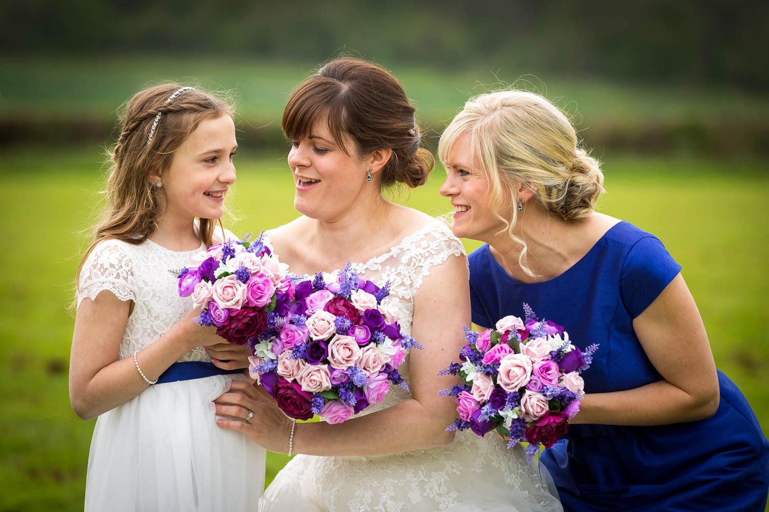 Three attendants in bridal party share joyful moment while holding vibrant purple and pink bouquets in outdoor setting.